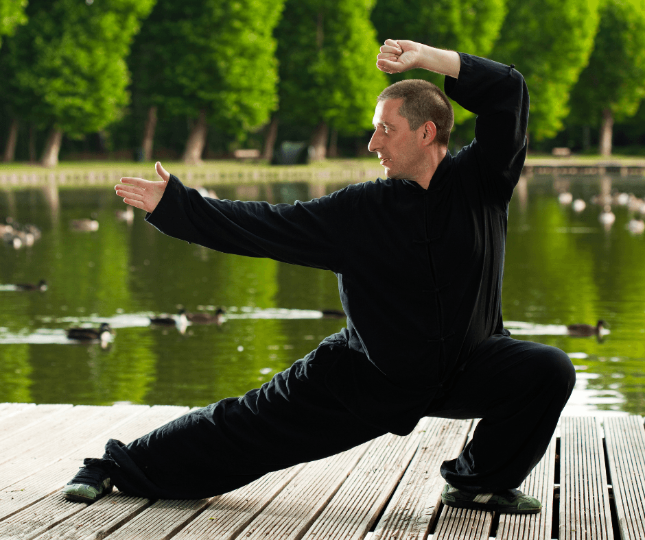 Mężczyzna w czarnym stroju ćwiczy tai chi na drewnianym pomoście nad jeziorem; stoi w pozycji bojowej z jedną ręką wyciągniętą do przodu, a drugą uniesioną nad głową. W tle widać wodę, drzewa i pływające kaczki.